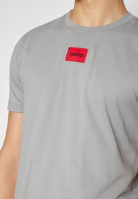 Hombre vistiendo una camiseta gris claro con un pequeño parche cuadrado rojo con "HUGO" impreso en negro en el pecho.