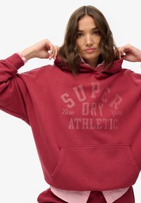 Czerwona, oversizeowa bluza z kapturem wykonana z miękkiego materiału, z kieszenią z przodu oraz napisem "SUPER DRY ATHLETIC". W komplecie z jasnoróżową koszulką pod spodem.