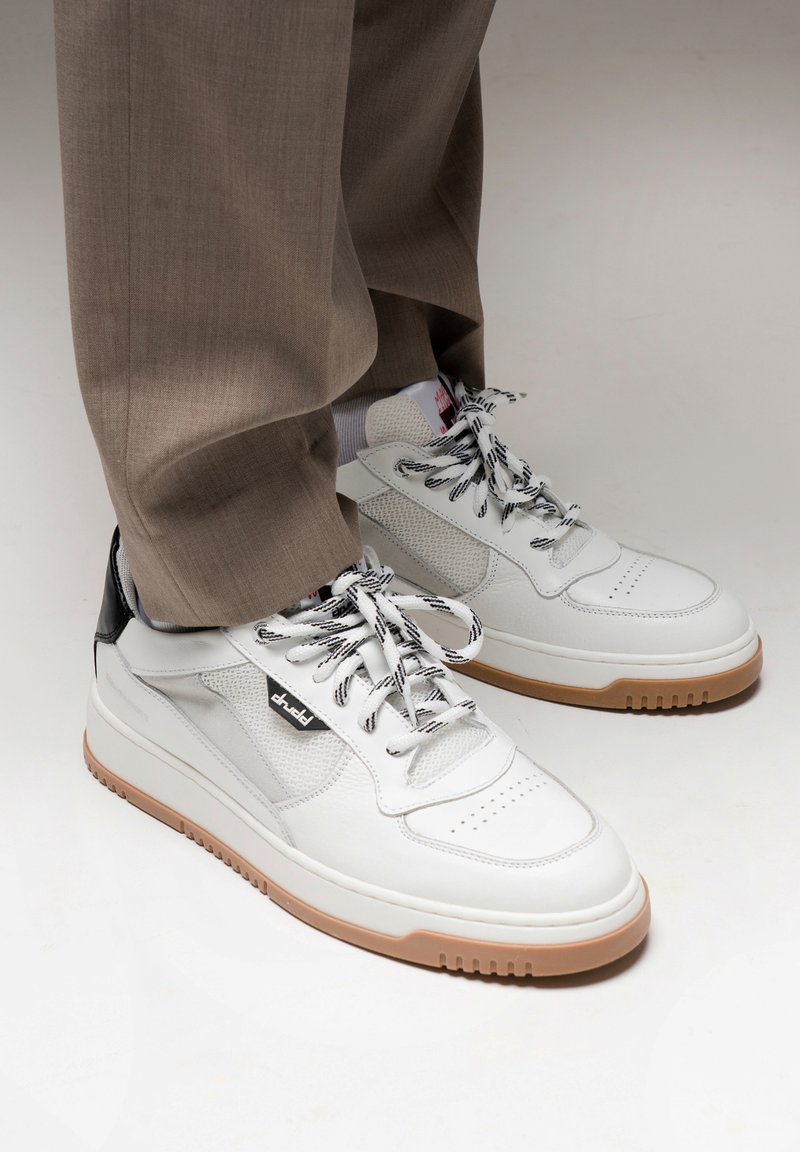 White leather sneakers with black accents, textured mesh panels, and black and white patterned laces. Gum rubber sole provides traction.