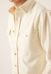 Cream-colored flannel shirt with a button-down collar, two front pockets, and brown buttons. Soft texture visible.