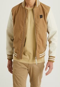 Brun bomberjacka med vita läderslevar, ribsöm i ärmarna och hög krage. Har en dragkedja framtill och en logotyp på bröstet.