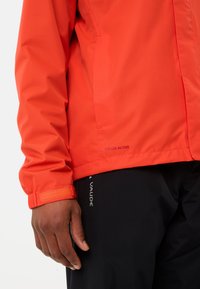 Rode waterdichte jas met een textuur oppervlak, elastische manchetten en "Ceplex Active" branding. Gecombineerd met zwarte broek met "Vaude" logo.