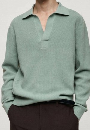 Person iført en lysegrøn strikket sweater med en bred krave og dyb V-udskæring, kombineret med mørkebrune bukser og et sort bælte.