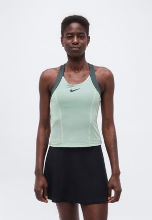 Kvinde iført en lysegrøn Nike tanktop og sort nederdel, stående med afslappede arme mod en ensfarvet hvid baggrund.