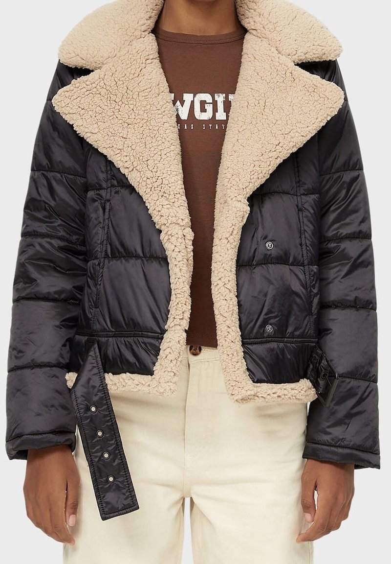 Giacca corta imbottita nera con collo e fodera in pelliccia sintetica beige, dettagli con bottoni e finitura strutturata, abbinata a una camicia marrone.