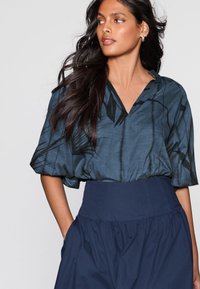 Mørkeblå bluse med korte ærmer og v-hals med sorte bladmotiver, kombineret med et højhalset, marineblåt skørt med rynkede detaljer.