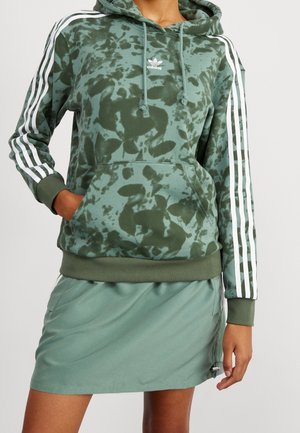 Frau trägt einen grünen Camouflage Adidas Hoodie mit weißen Streifen an den Ärmeln und einem einfarbigen grünen Rock, Hände in den vorderen Taschen.