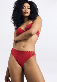 Sostén de encaje rojo con borde festoneado y aros, combinado con un tanga de encaje rojo a juego. Ambos tienen una pequeña etiqueta de marca en el lado.