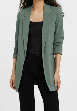 Femme portant un blazer vert sauge avec les manches retroussées sur un haut noir et un pantalon noir, sur un fond clair uni.