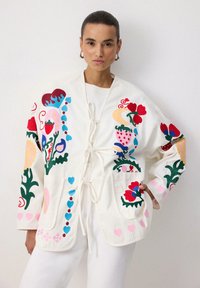 Valge, tikandiga jakk, millel on lillemustrid punases, roosas, sinises ja rohelises. Lõdva lõikega, vöökoha seotav ja kahe esisegmendiga taskud.