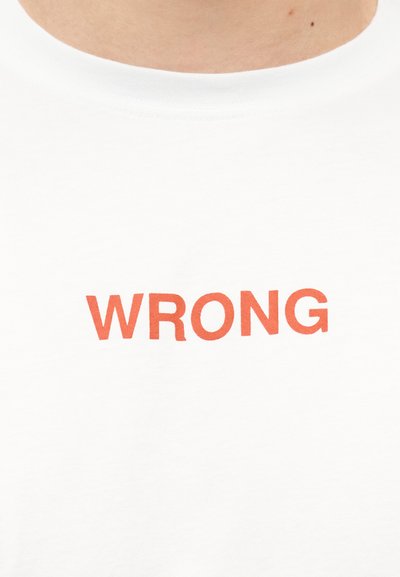 Gros plan sur une chemise blanche avec le mot "WRONG" imprimé en lettres rouges grasses sur la poitrine.