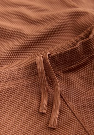 Brauner, strukturierten Stoff mit Wabenmuster, versehen mit einem Kordelverschluss und glatten Kanten. Das Material wirkt weich und flexibel.