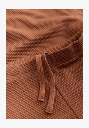 Pruun tekstuuriga kangas mesilasmustri mustriga, millel on paelaga kinnitussüsteem ja siledad servad. Materjal tundub pehme ja paindlik.