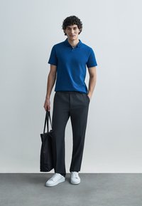 Μπλε polo μπλουζάκι με κοντά μανίκια, σε άνετη εφαρμογή, συνδυασμένο με σκούρο στενό παντελόνι και λευκά αθλητικά παπούτσια. Το μοντέλο κρατά μια μαύρη τσάντα tote.