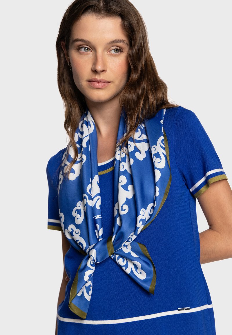 Haut en maille bleu avec des accents blancs et dorés, présentant un foulard bleu à motifs noué au cou. Texture lisse, manches courtes et coupe ajustée.