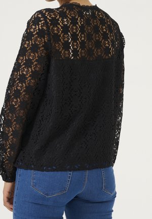 Personne portant un haut en dentelle noire à manches longues à motifs floraux sur un tissu uni, associé à un jean en denim bleu, vue de dos.