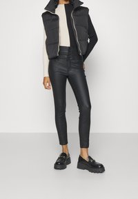 Svart pufferväst över en figursydd krämfärgad ribbad tröja, kombinerad med högmidjade svarta läderliknande byxor och kraftiga svarta loafers med gulddetaljer.