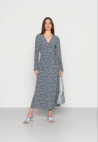 Vestido florido envolvente em azul-marinho com pequenas flores brancas, mangas longas e decote em V. Combinado com sandálias de salto bloco azul-claro e uma mala a condizer.