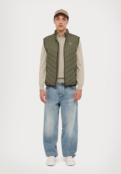 Ελαιόχρωμο καπιτονέ γιλέκο πάνω από μια μπεζ μπλούζα με μακριά μανίκια, συνδυασμένο με φαρδιά ανοιχτό μπλε τζιν και λευκά αθλητικά παπούτσια. Διαθέτει ράφι με λογότυπο.