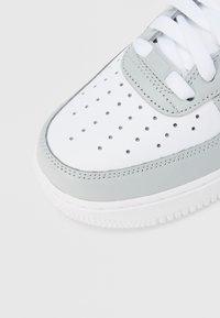 Zbliżenie na czubek białego trampka z szarą lamówką, otworami perforowanymi i białymi sznurowadłami na białym tle.