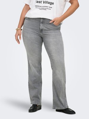 Persona che indossa jeans grigi slavati a gamba dritta, scarpe slip-on nere, maglietta bianca con grafica e braccialetto d'oro, in piedi con una mano in tasca.