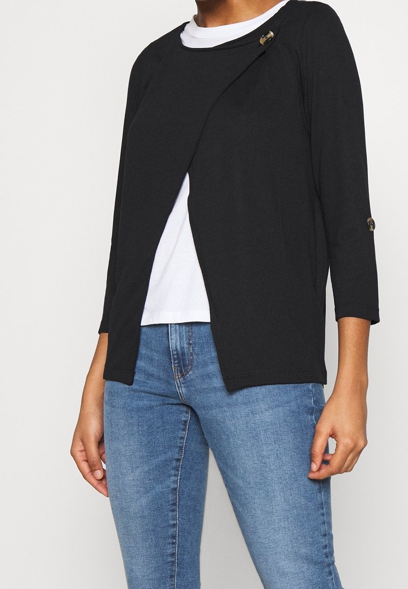 Personne portant un cardigan noir à boutons sur une chemise blanche et un jean bleu, debout devant un fond uni.