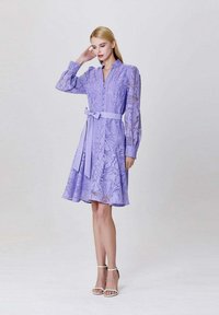 Robe lavande à manches longues et ceinture à nouer. Présente des broderies florales et une jupe évasée. Portée avec des talons blancs à lanières.