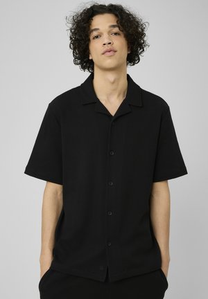 Jeune personne aux cheveux bouclés portant une chemise noire à manches courtes boutonnée, debout devant un fond gris uni.
