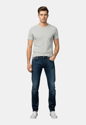 Giovane uomo in piedi, indossa una maglietta grigio chiaro, jeans blu scuro, scarpe da ginnastica bianche, con una mano in tasca davanti a uno sfondo semplice.