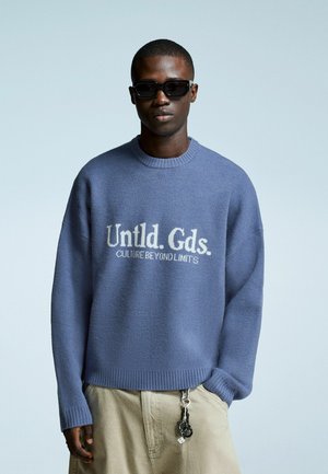 Young man wearing dark sunglasses, oversized blue sweater with "Untld. Gds. CULTURE BEYOND LIMITS" text, beige pants, and keychain hanging.