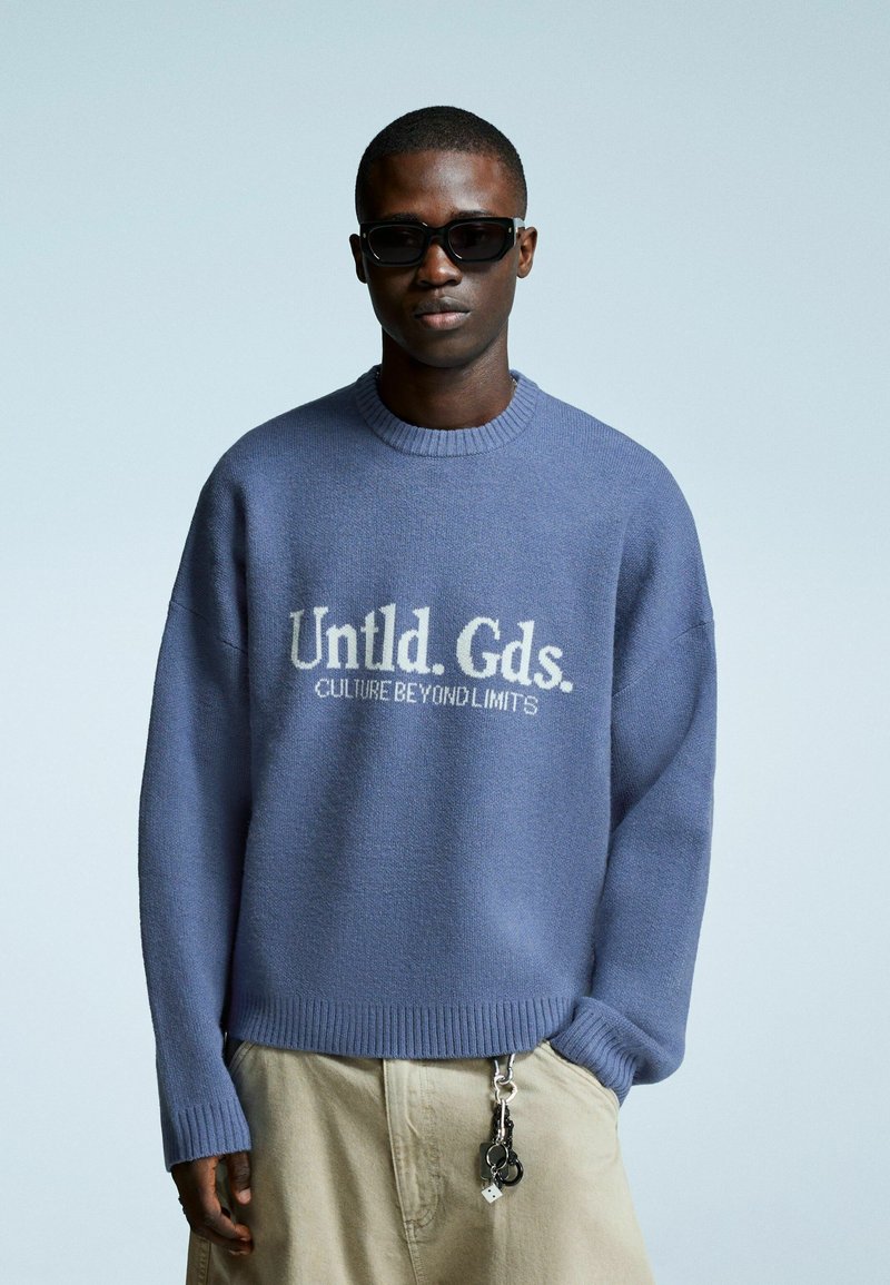 Jongeman met donkere zonnebril, oversized blauwe trui met de tekst "Untld. Gds. CULTURE BEYOND LIMITS", beige broek en een sleutelhanger die hangt.