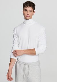 Biała, długoucha, z golfem, wykonana z miękkiego materiału, z dopasowanymi rękawami i bezszwowym wykończeniem, w połączeniu z jasnoszarymi spodniami dresowymi ze sznurkiem.