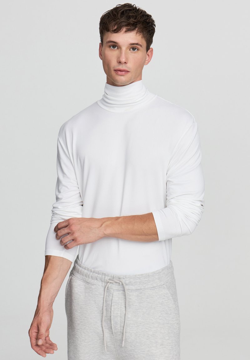 Biała, długoucha, z golfem, wykonana z miękkiego materiału, z dopasowanymi rękawami i bezszwowym wykończeniem, w połączeniu z jasnoszarymi spodniami dresowymi ze sznurkiem.