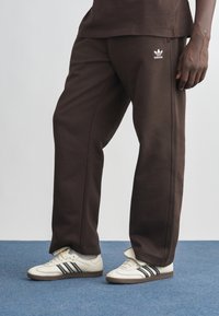 Barna, pamut melegítőnadrág, karcsúsított fazon, fehér Adidas logóval. Krémszínű cipőkkel, fekete csíkkal párosítva. Kék szőnyeg hátterével.