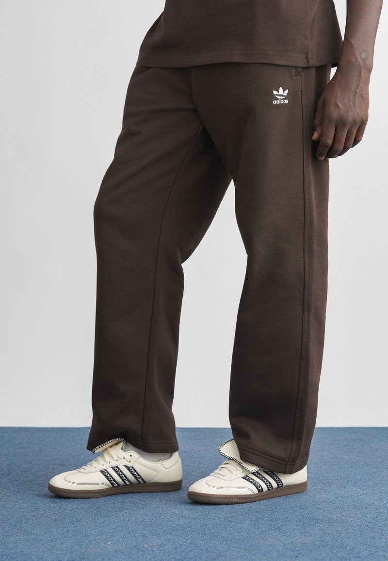 Barna, pamut melegítőnadrág, karcsúsított fazon, fehér Adidas logóval. Krémszínű cipőkkel, fekete csíkkal párosítva. Kék szőnyeg hátterével.