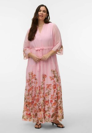 Femeie îmbrăcată într-o rochie lungă roz cu flori, mâneci transparente și o centură legată în talie, stând în fața unui fundal alb simplu.