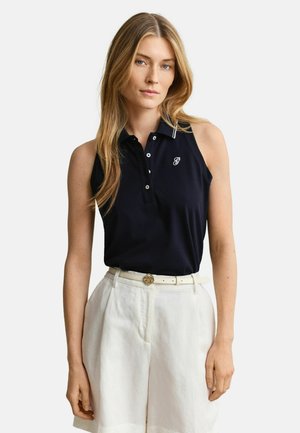 Femme aux longs cheveux blonds portant un polo noir sans manches et un short blanc taille haute avec une ceinture blanche.