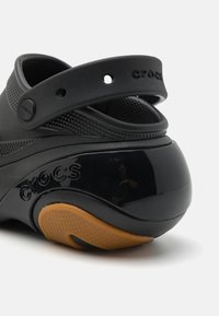 Svarta Crocs med en strukturerad ovandel, ett bakre rem och en glänsande svart klack. Sulan har ett mönstrat gummimaterial i tan för bra grepp.