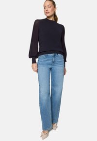 Femme portant un pull bleu marine foncé avec des manches bouffantes texturées, un jean droit bleu clair et des chaussures pointues beiges avec des nœuds.