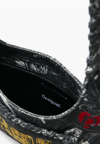 Sort mønstret håndtaske med lynlåsåbning, der viser en "Desigual"-etiket indeni og en flettet håndtagsdetalje.