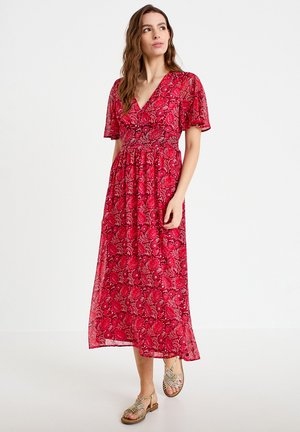 Kvinde med langt hår iført en rød blomstret V-hals midi-kjole og beige flettede sandaler, stående mod en hvid baggrund.