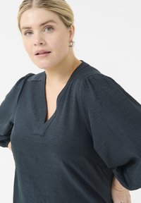Donkerblauwe, gestructureerde top met een V-hals en pofmouwen. De stof heeft een subtiele glans, met een relaxed fit en strakke lijnen.