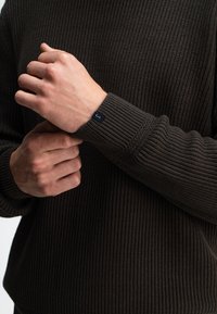 Dunkelbrauner gerippter Strickpullover mit rundem Halsausschnitt und engen Bündchen. Verfügt über ein kleines schwarzes Etikett am Handgelenk. Deutliche Struktur erkennbar.