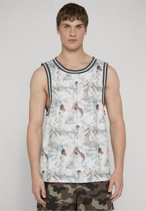 Ung mand iført en ærmeløs skjorte med abstrakt mønster og sorte kanter samt camouflage cargoshorts, stående mod en ensfarvet baggrund.