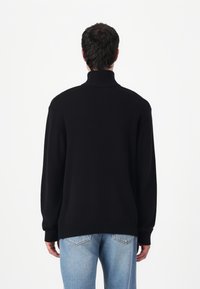 Maglione nero a collo alto in maglia con polsini e orlo a coste, caratterizzato da una vestibilità rilassata e una superficie texture. Indossato con jeans in denim azzurro chiaro.