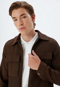Chaqueta marrón acanalada con cremallera, dos bolsillos frontales y un cuello, combinada con una camiseta polo blanca con tapeta de botones.