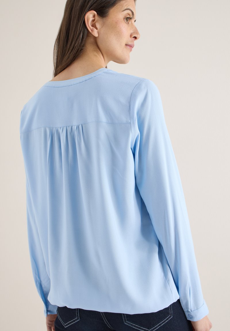 Blouse à manches longues bleu clair avec un col rond, présentant des plis au dos et des poignets boutonnés. Texture lisse et coupe ample.