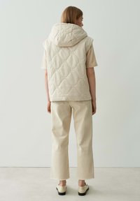 Beige gesteppte Weste mit Kapuze, die eine weiche Textur aufweist. Über einem hellbeigen Hemd getragen, kombiniert mit cremefarbenen, hüftlangen Hosen.