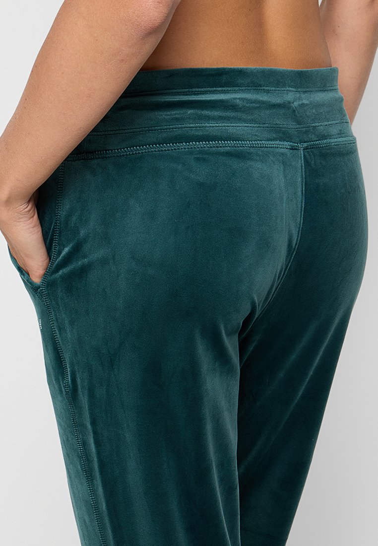 Osoba w ciemnozielonych welurowych spodniach z widocznymi szwami, z ręką w bocznej kieszeni, pokazana od talii do połowy uda.