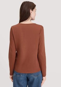 Vrouw met lang bruin haar, gekleed in een effen bruin langmouwen shirt en blauwe spijkerbroek, gezien vanaf de achterkant tegen een lichte achtergrond.